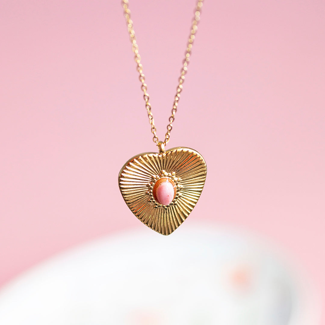 Image shows a gold plated sunburt heart necklace with a pink gemstone at the centre displayed on a pink background