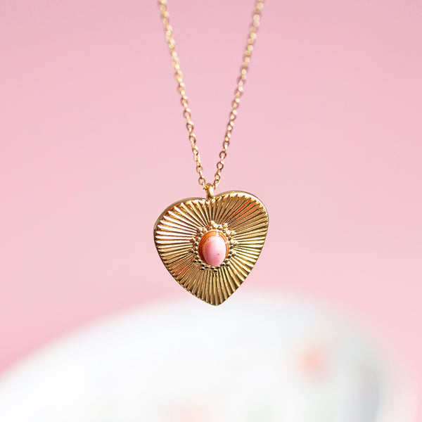 Image shows a gold plated sunburt heart necklace with a pink gemstone at the centre displayed on a pink background