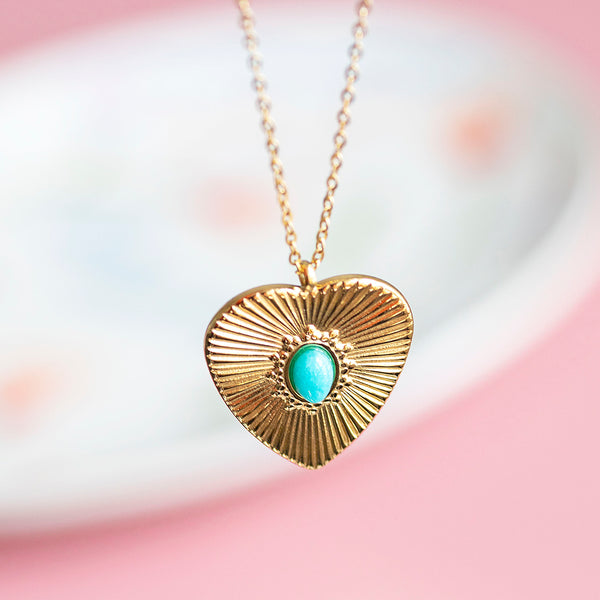 Image shows a gold plated sunburtst heart necklace with turquoise stone detail with a pink background