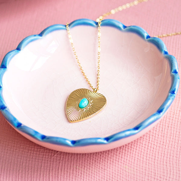 Image shows a gold plated sunburtst heart necklace with turquoise stone detail in a scalloped trinket dish