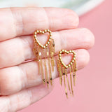 Image shows a hand holding a pair of gold plated dotted heart earrings with chain drop detail