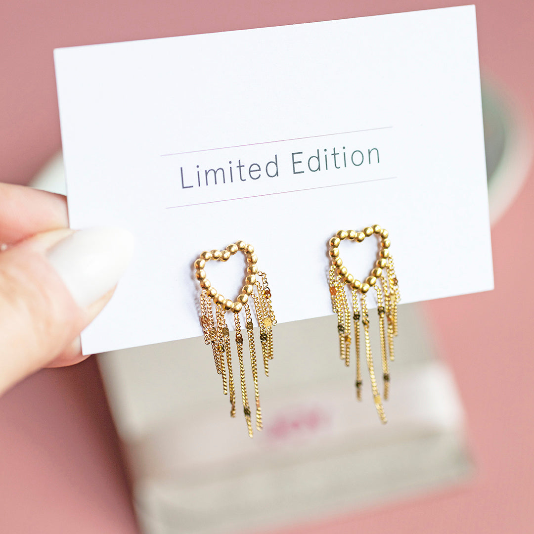 Image shows a hand holding a pair of gold plated dotted heart earrings with chain drop detail mounted on a limited edition card