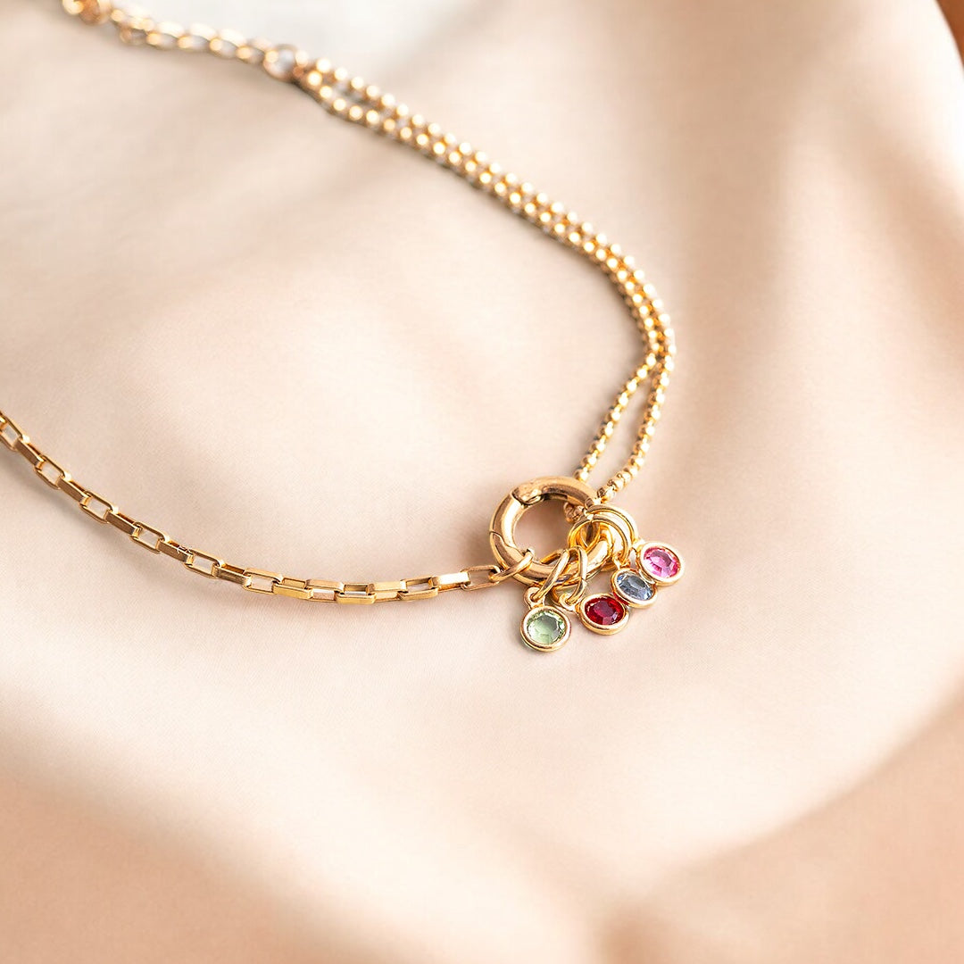 Image shows a multi birthstone gold charm bracelet on a neutral satin fabric background