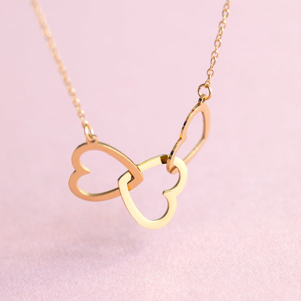 Image shows a gold plated trio heart necklace on a pink background
