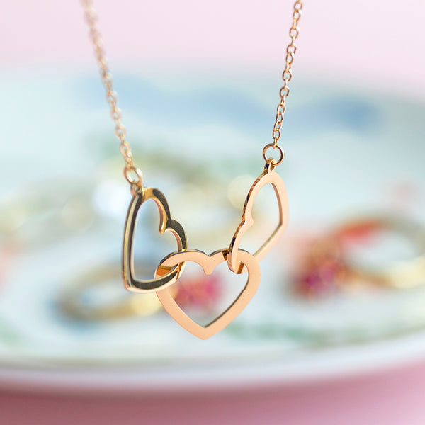 Image shows a gold plated trio heart necklace on a pastel background