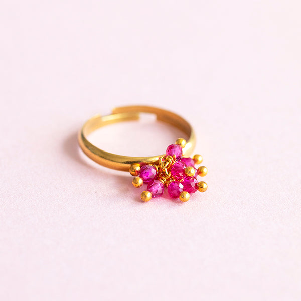 Image show a gold plated adjustable ring with a cluster of ruby gemsone charms on a pink background