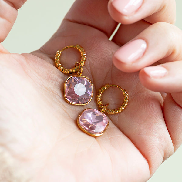 Image shows a womans hand holding a pair of gold hopp earrings with pink crystal charms