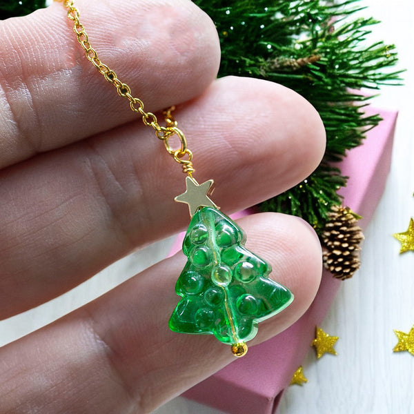 image shows a hand holding green glass Christmas tree necklace with gold star detail on a pink festive background