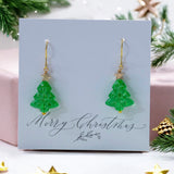 Image shows Christmas tree earrings on a merry Christmas card against a pink festive themed background