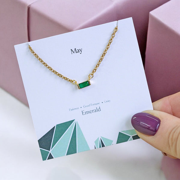 image shows a hand holding a birthstone characteristics card with a gold may emerald birthstone baguette necklace