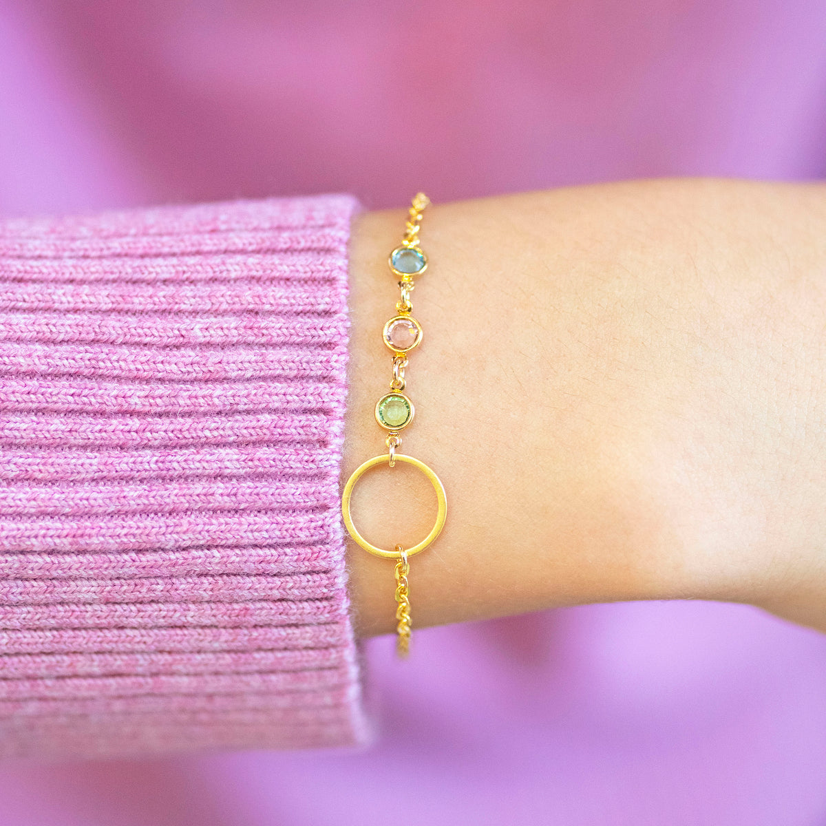 Gold plated Mother and Three Children Birthstone Bracelet with open circle charm and three stones in blue, pink and green, styled with a pink jumper.