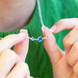 Close-up of hands holding a delicate silver necklace with blue and pink birthstones against a green background.