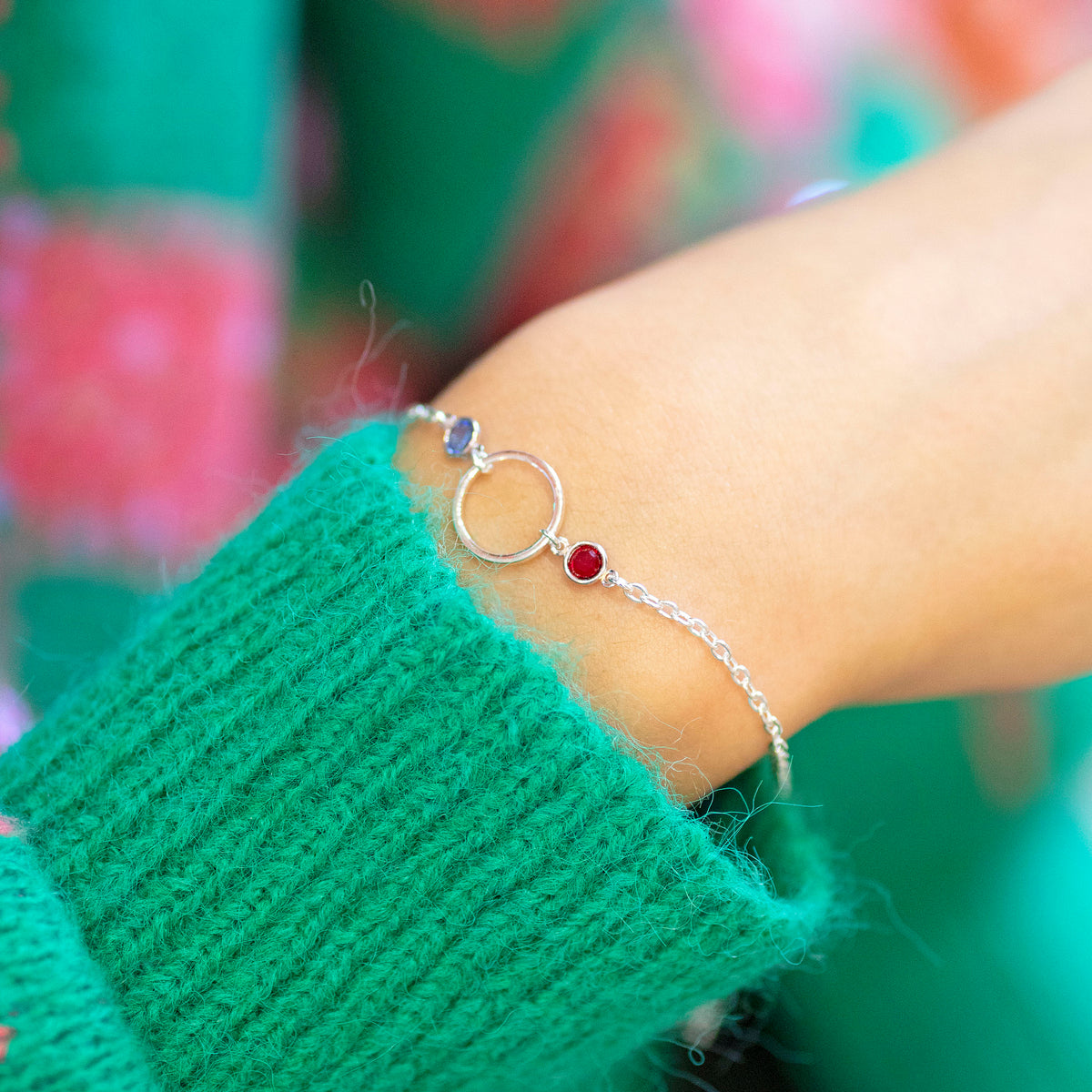 Silver plated Mother and Two Children Birthstone Bracelet with open circle charm and sapphire and garnet birthstones, styled with a green knit jumper.