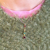 Gold necklace with colourful birthstones worn by a person against a textured green background