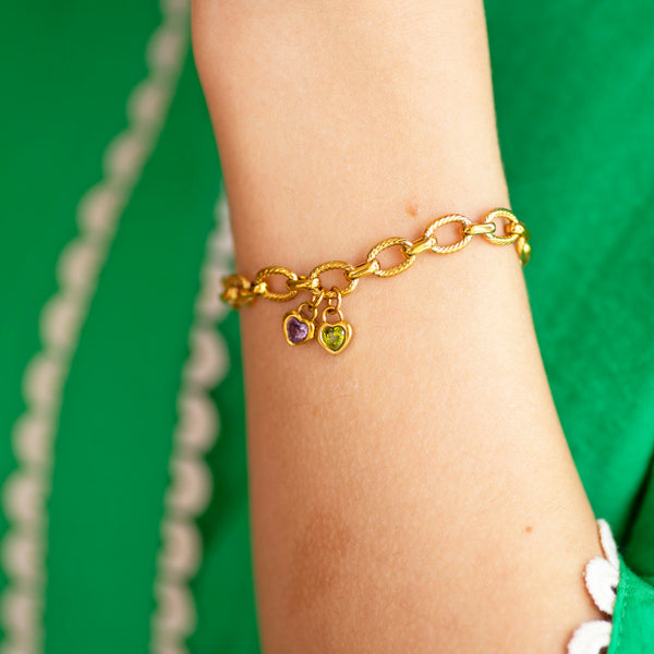 Gold charm chain bracelet with heart shaped birthstones on a wrist against a green background