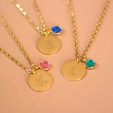 Gold modern initial birthstone disc necklaces with engraved letters E, K, and L, each paired with a colourful birthstone charm on a warm neutral background