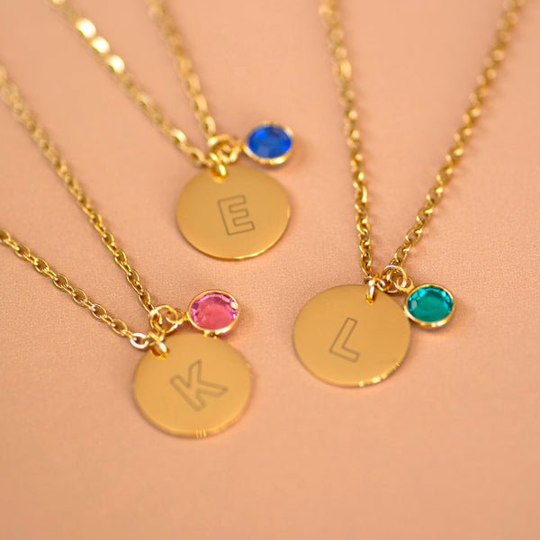 Gold modern initial birthstone disc necklaces with engraved letters E, K, and L, each paired with a colourful birthstone charm on a warm neutral background