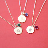 Silver modern initial birthstone disc necklaces with engraved letters A, C and S, each paired with a coloured birthstone charm on a soft coral background.