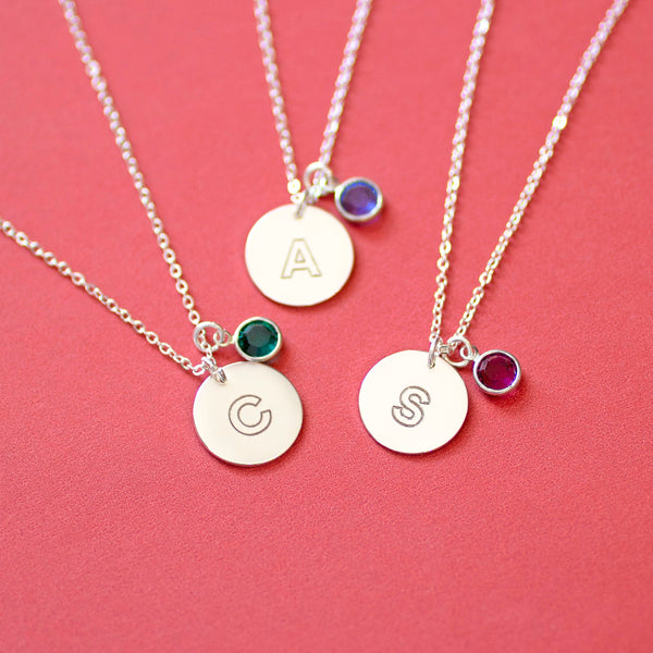 Silver modern initial birthstone disc necklaces with engraved letters A, C and S, each paired with a coloured birthstone charm on a soft coral background.
