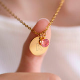 Close up of gold initial birthstone necklace with engraved letter K and pink birthstone charm, held to highlight fine detail and shine.