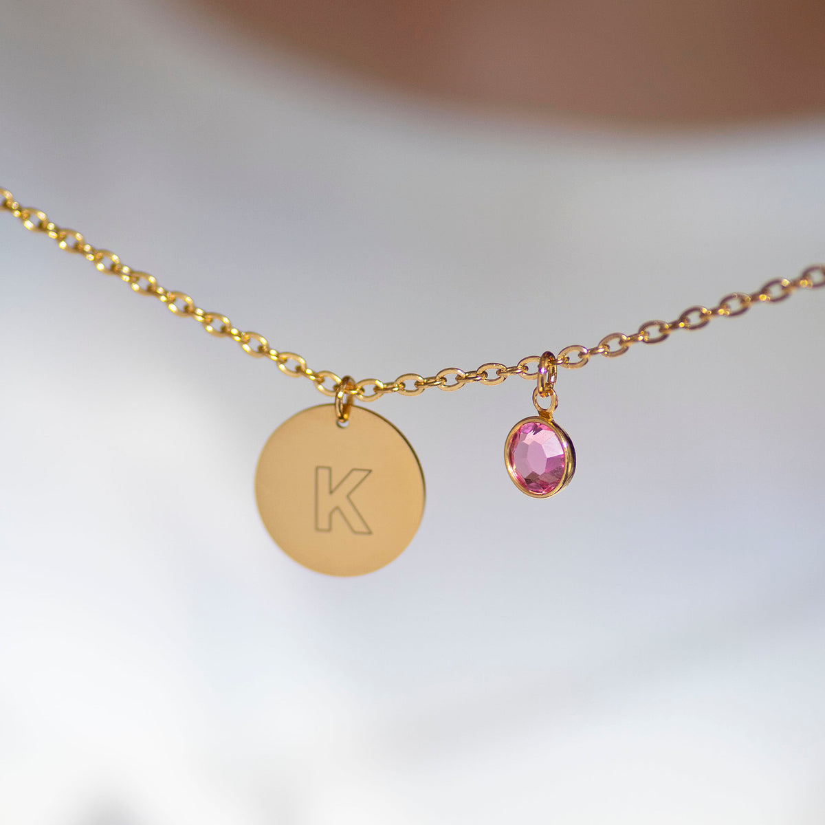 Minimal gold initial disc necklace with engraved K and pink birthstone charm on a fine chain against a soft blurred background.