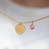 Minimal gold initial disc necklace with engraved K and pink birthstone charm on a fine chain against a soft blurred background.