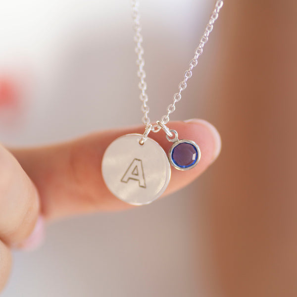 Close up of silver modern initial birthstone disc necklace with engraved letter A and blue birthstone charm, held to highlight fine detail and shine.