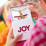 Personalised Week Of Charms Necklace in gold presented in JOY by Corrine Smith gift box with interchangeable moon, heart, star and horseshoe charms against a colourful background.