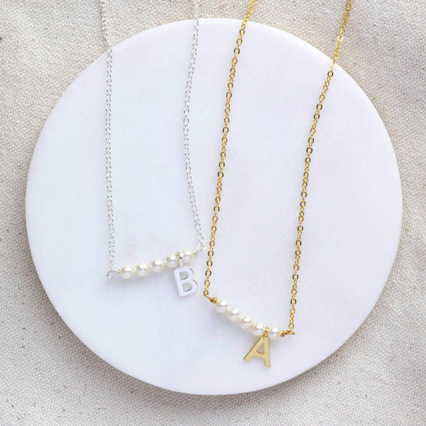 Two asymetric pearl bar initial necklaces silver with the initial B and Gold with the initial A lying a white circle coaster