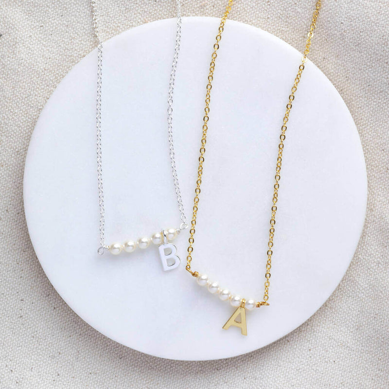 Two asymetric pearl bar initial necklaces silver with the initial B and Gold with the initial A lying a white circle coaster