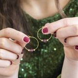 Image shows model holding gold beaded birthstone hoop earrings with February birthstone