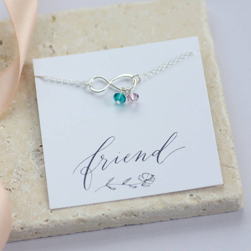 Best friend infinity birthstone charm bracelet with blue zircon and light amethyst present on a friend sentiment card lying on a cream stone tile