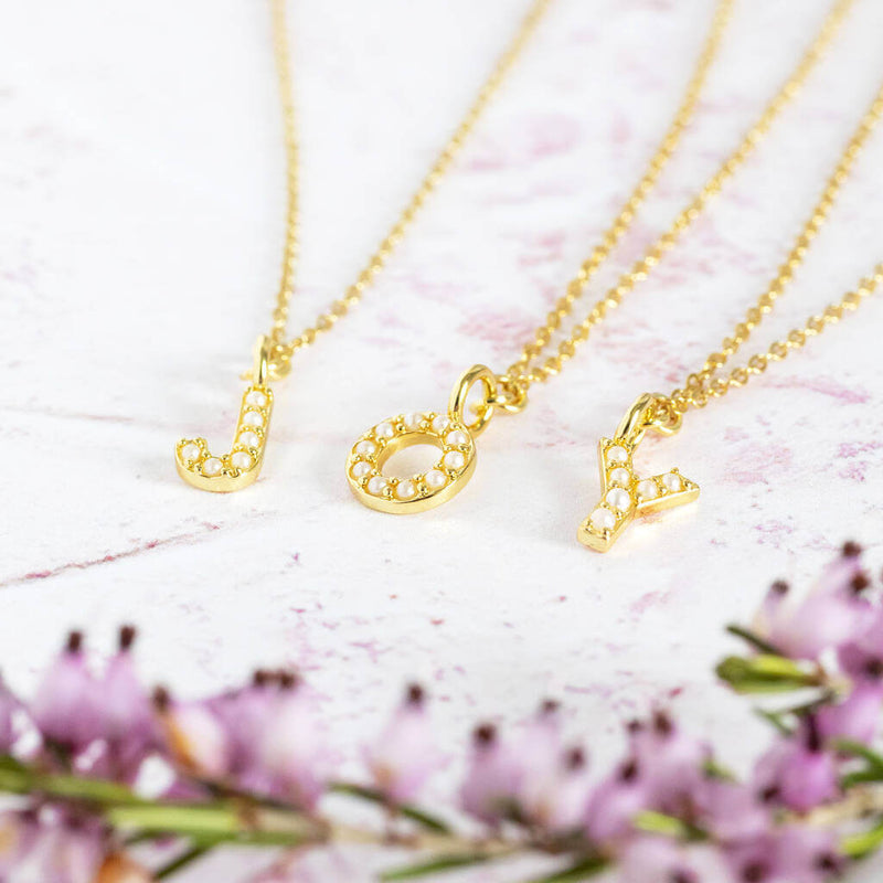 Three gold dainty pearl necklaces with the initials J O Y lying on a pinky white background with pink flowers blurred out
