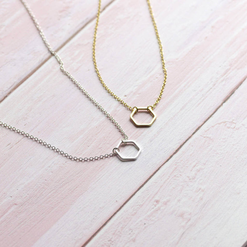 Image shows from left to right: silver plated hexagon outline necklace and gold plated hexagon outline necklace on a pale pink backdrop.