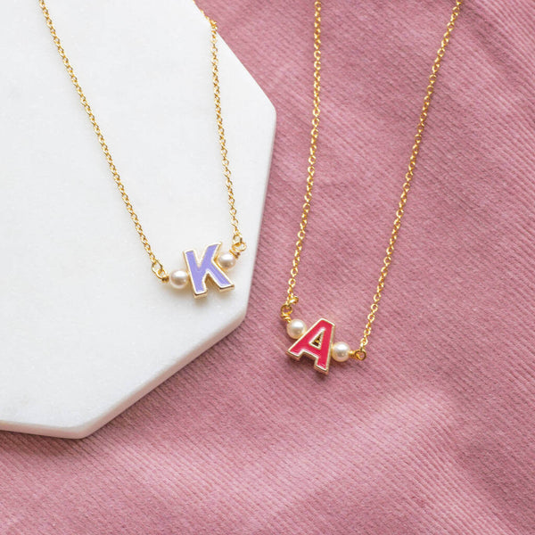 Two enamel initial necklaces with pearl details one with a K initial sitting a white hexagon coaster and one with A initial sitting on pink cord 