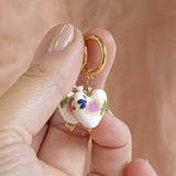Image shows model holding Floral Porcelain Heart Huggie Earrings