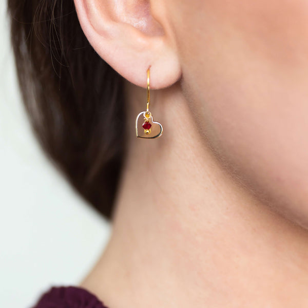 Image shows model wearing gold heart birthstone charm earring with July birthstone