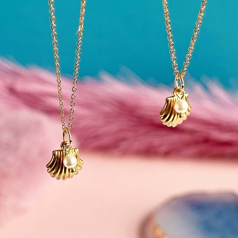 Image shows two gold plated shell necklaces with pearl detail hanging in front of a pink and blue backdrop