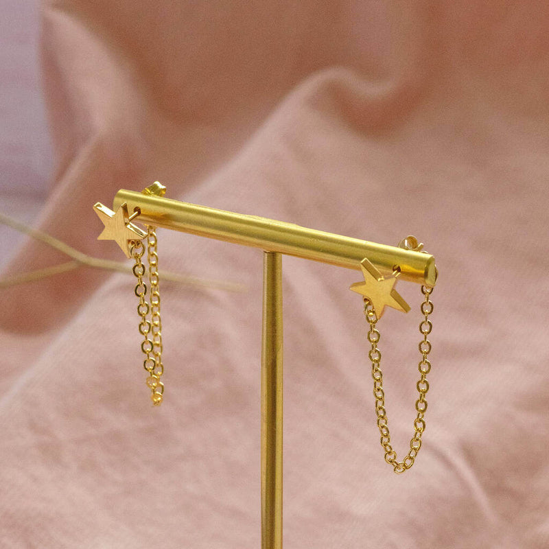 Image shows Gold Plated Star Earrings with Chain Drop Detail hanging on a gold earring stand