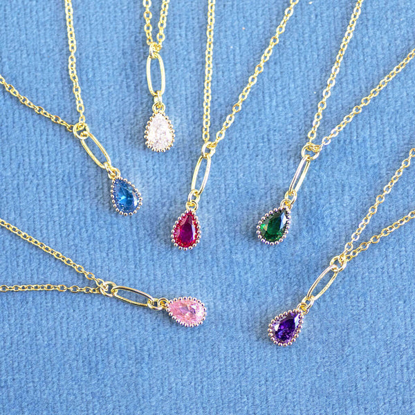 Image shows a selection of Gold Plated Teardrop Birthstone Pendant Necklace lying on blue material