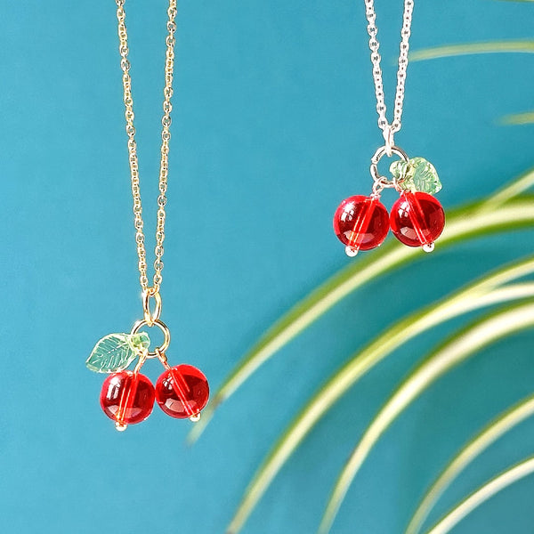 Image shows one gold and one silver plated glass cherry necklace hanging in front of a blue backdrop