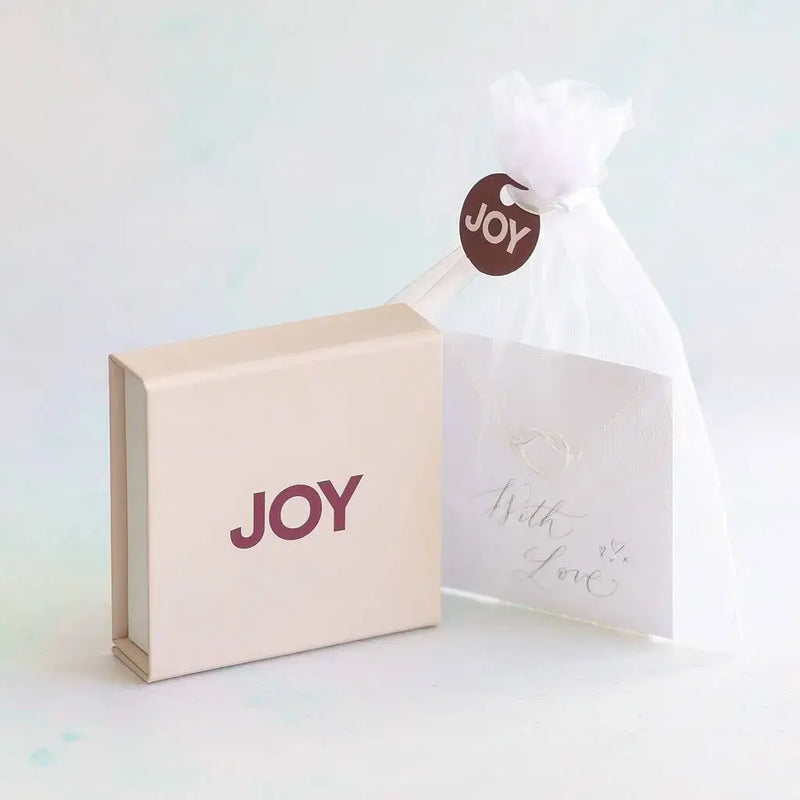 Image shows JOY gift box with JOY logo in maroon, and white organza bag with a sentiment card inside, finished with a white ribbon and a JOY tag