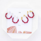 Image shows silver and gold July Birthday Ruby Birthstone Beaded Huggie Earrings lying on a July birthstone characteristic card