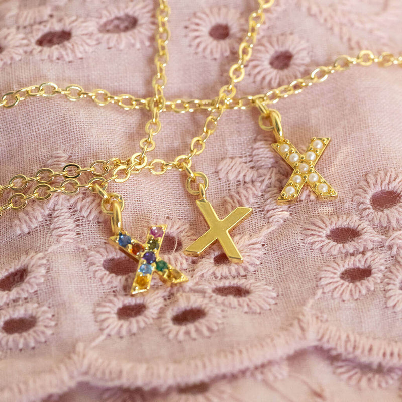 Three pearl kiss 'X' charm bracelet lying on pink material  one with rainbow X one with gold X. and one with pearl X