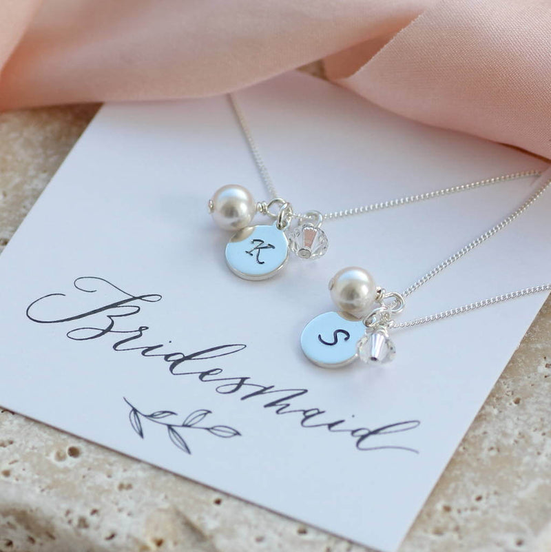 Two personalised bridesmaid charm necklaces lying on a bridesmaid sentiment card one with the K initial and one with a S