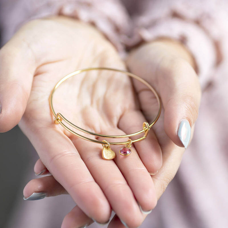 Image shows model holding personalised heart of gold birthstone bangle with A engraved on the heart and light amethyst birthstone on the palms of her hand