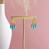 Image shows Triangle Studs with Turquoise Bead Drop Detail on a gold earring stand