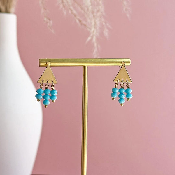 Image shows Triangle Studs with Turquoise Bead Drop Detail on a gold earring stand