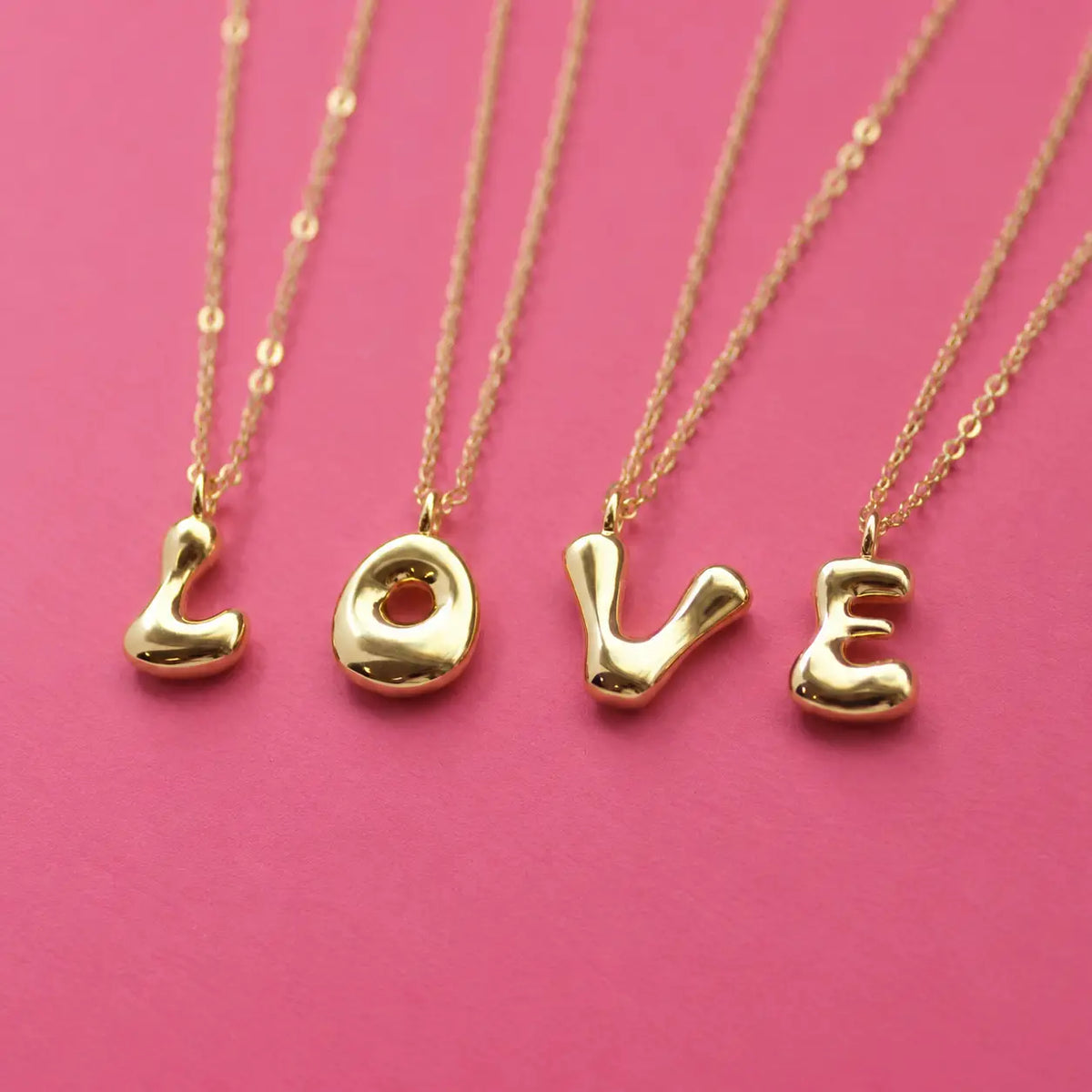Personalised gold bubble initial necklaces spelling LOVE with chunky letter pendants on a pink background
