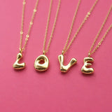 Personalised gold bubble initial necklaces spelling LOVE with chunky letter pendants on a pink background